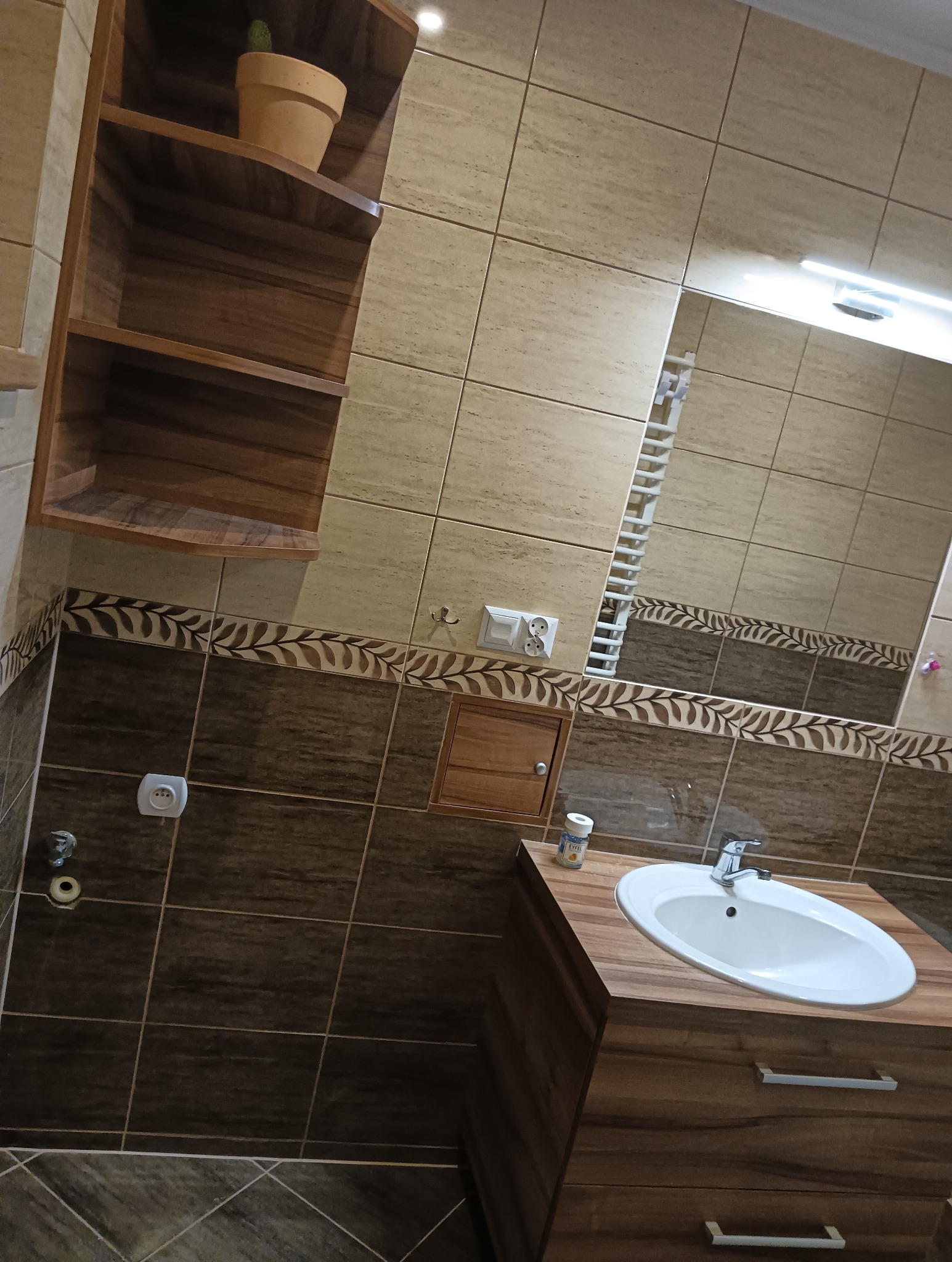 Ukośne ujęcie wykończonej łazienki z brązowo-beżowymi płytkami, drewnianą szafką pod umywalką, otwartą półką z kaktusem i dekoracyjnym pasem z motywem liści.