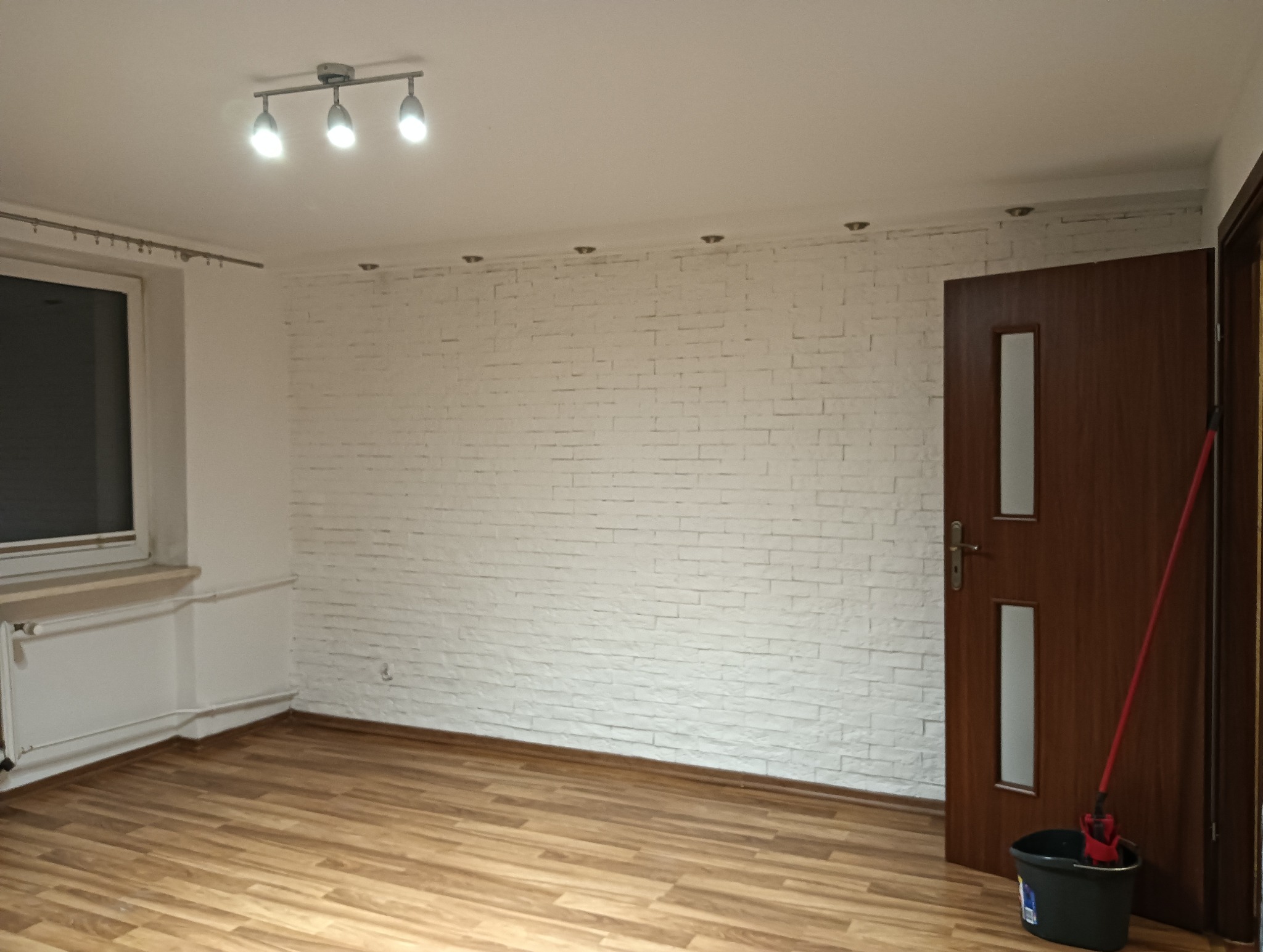 Jasne wnętrze pokoju z drewnianą podłogą, białą ceglaną ścianą i brązowymi drzwiami, widoczny mop w wiadrze.