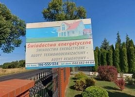 Tablica reklamowa z informacją o usługach: świadectwa energetyczne, audyt termomodernizacyjny, audyt remontowy, z ilustracją domu z czerwonym dachem, umieszczona na ogrodzeniu z cegły klinkierowej.
