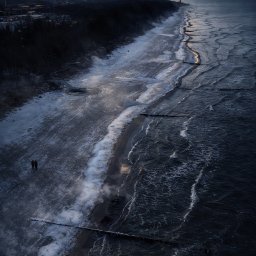 DRONOZA Studio - Widok z góry na zimową plażę z parą spacerującą wzdłuż brzegu, w oddali latarnia morska oświetlona zachodzącym słońcem, ciemne chmury na niebie.