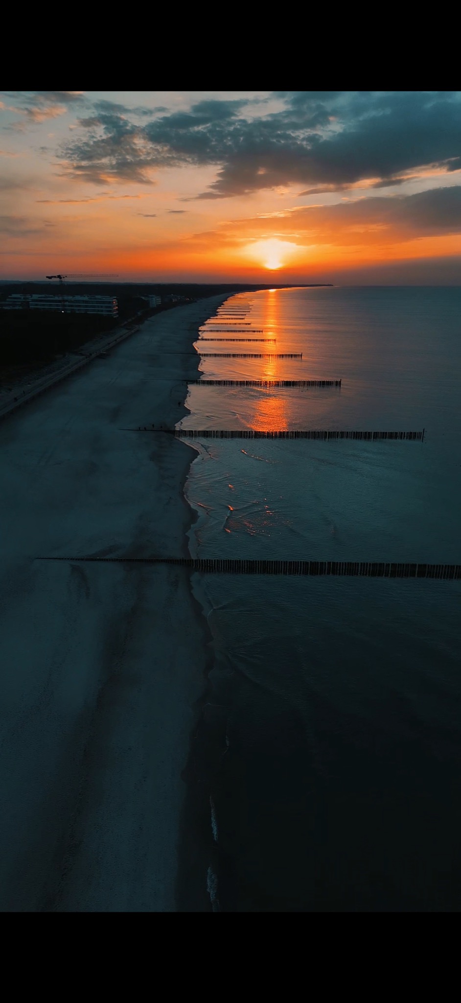 Widok z lotu ptaka na plażę o zmierzchu, z drewnianymi falochronami wzdłuż brzegu, odbijającymi pomarańczowe światło zachodzącego słońca.
