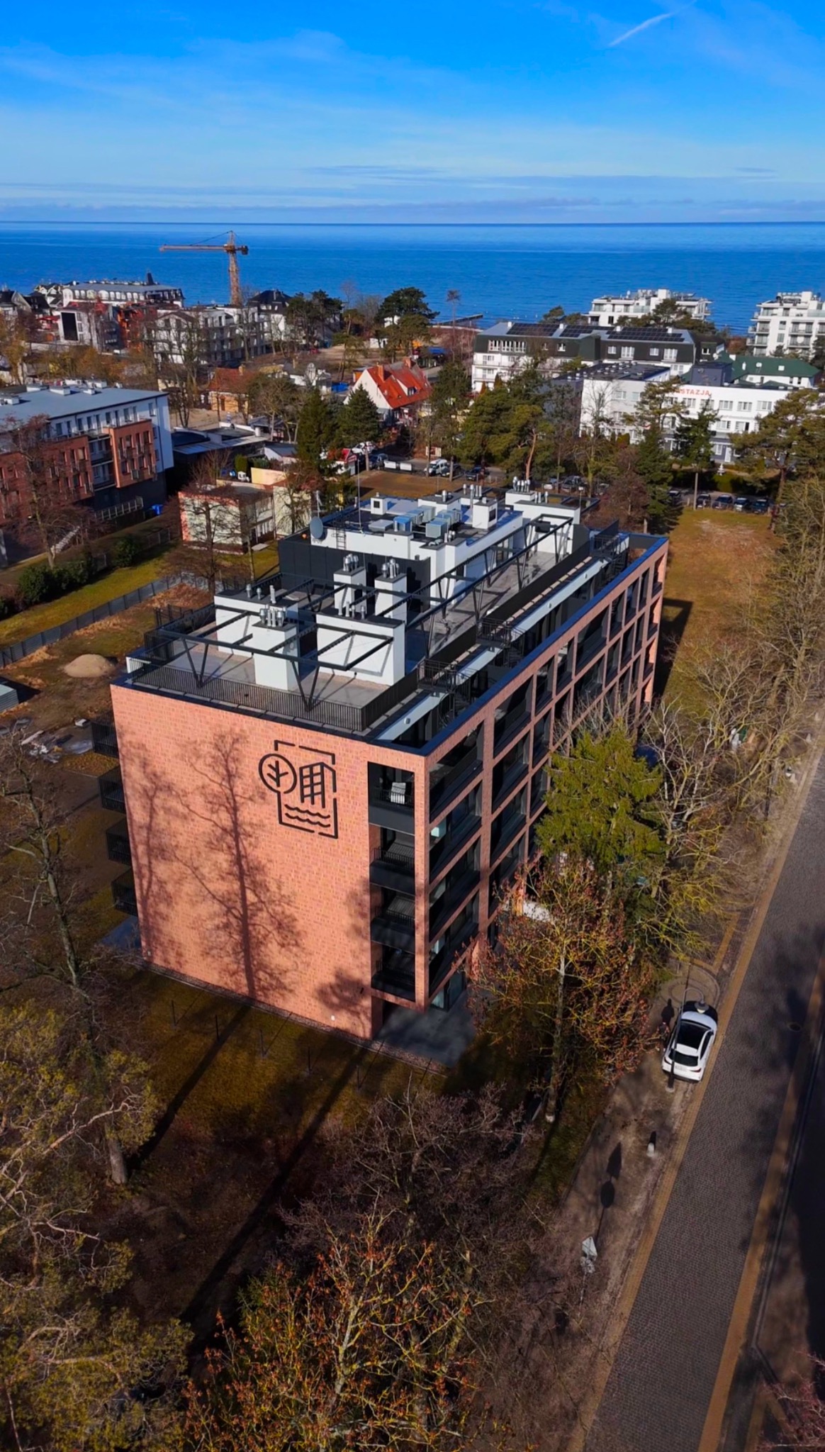 Widok z drona na nowoczesny, ceglany budynek apartamentowy z logo, z balkonami i tarasem na dachu, na tle morza i zabudowy miejskiej w słoneczny dzień.