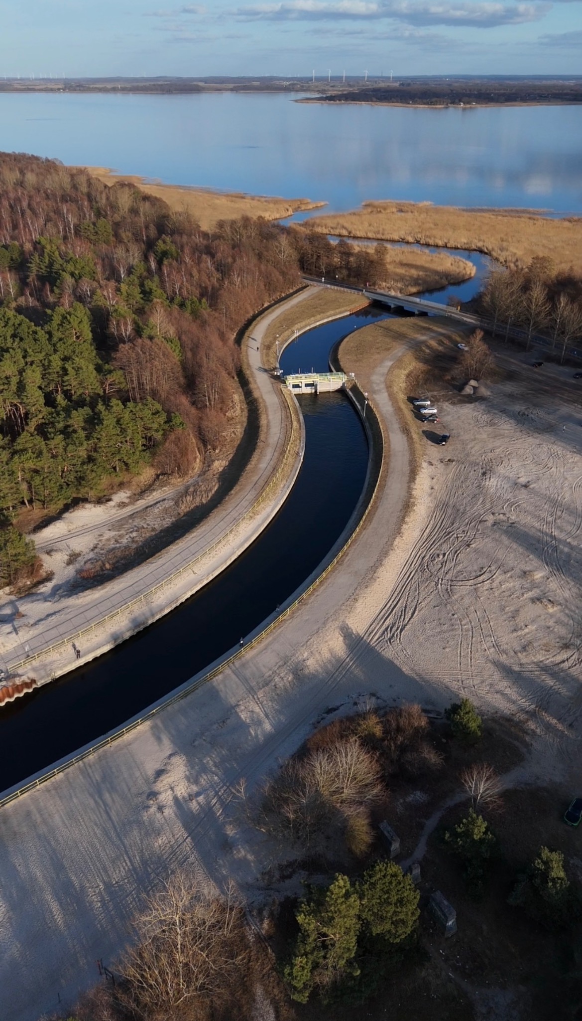 Widok z drona na kanał wodny z betonowymi brzegami, przecinający piaszczysty teren i łączący się z jeziorem. W tle widoczne turbiny wiatrowe.