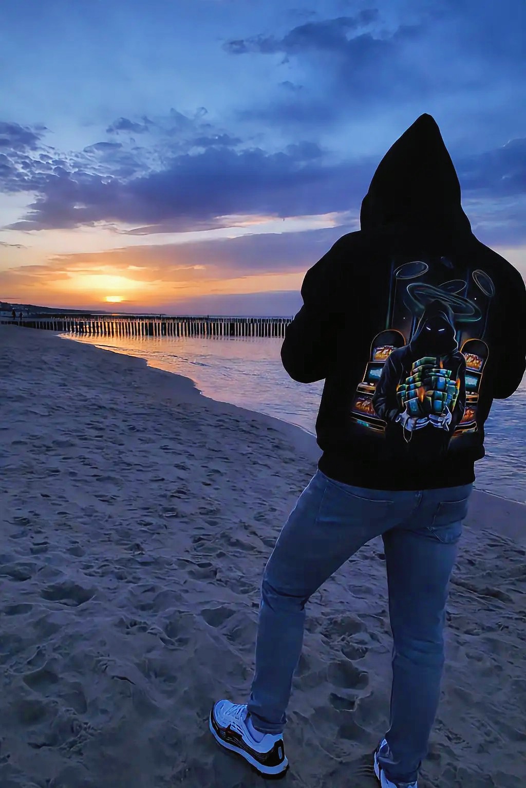 Osoba w bluzie z kapturem stoi tyłem na plaży o zmierzchu, obserwując zachód słońca nad morzem. Na bluzie widoczny graficzny wzór.
