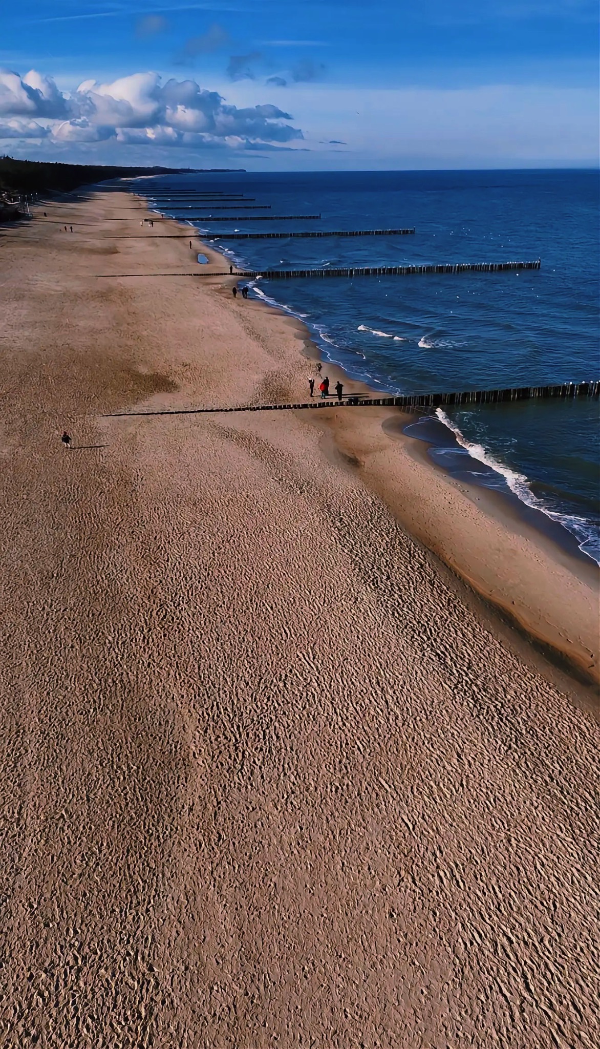 Szeroki kadr z lotu ptaka na piaszczystą plażę z drewnianymi falochronami wzdłuż brzegu, błękitne morze i niebo z chmurami w tle. Widoczni spacerujący ludzie.
