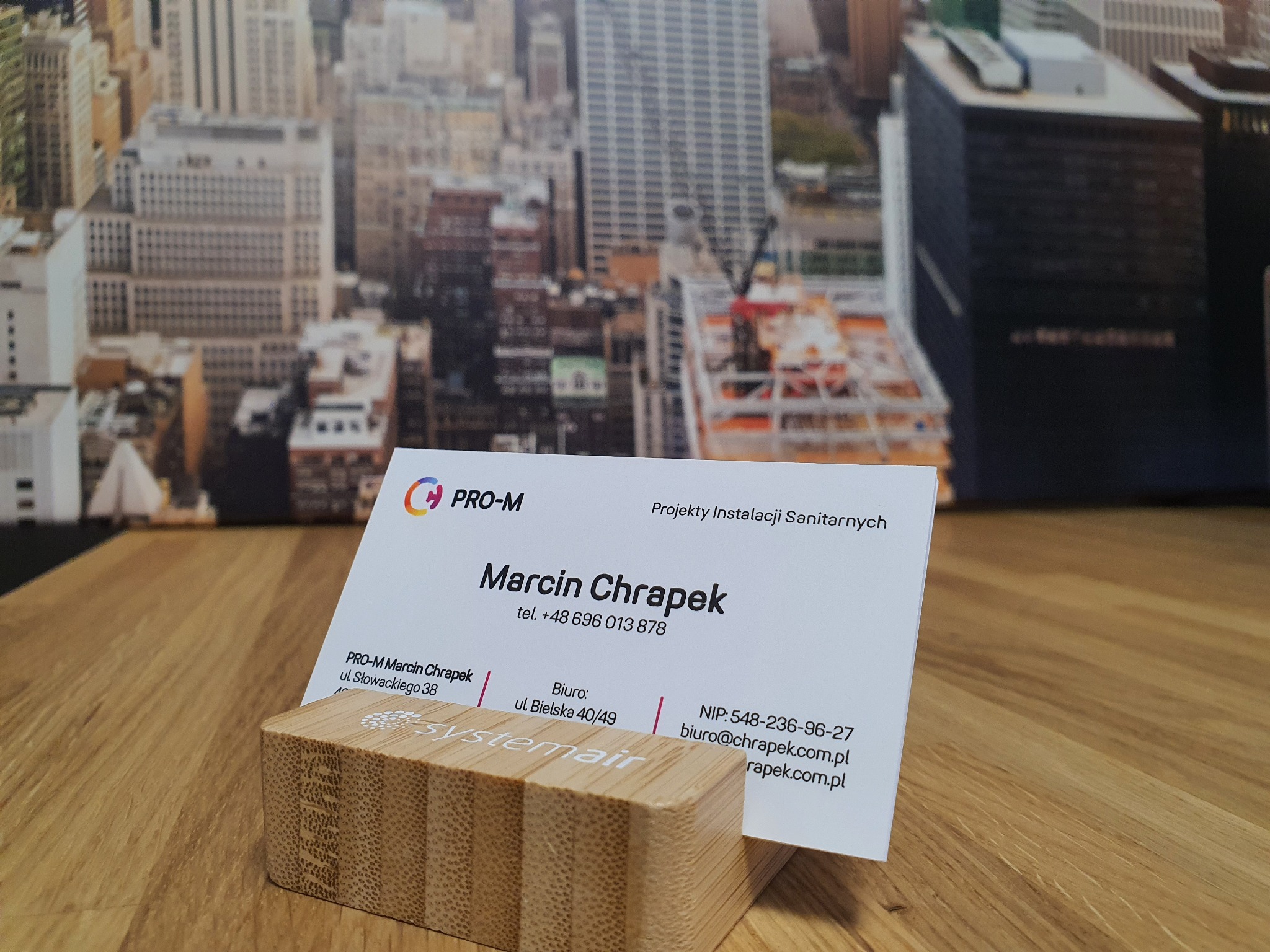 Wizytówka firmy PRO-M Marcin Chrapek, specjalizującej się w projektach instalacji sanitarnych, umieszczona w drewnianym stojaku na tle panoramy miasta.