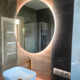 SZKŁO & ALU - Nowoczesna łazienka z okrągłym lustrem podświetlanym LED, białą umywalką nablatową i złotą armaturą na tle płytek imitujących drewno i marmur.