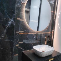 SZKŁO & ALU - Nowoczesna łazienka z umywalką nablatową z marmuru, okrągłym lustrem z podświetleniem LED i czarnymi płytkami imitującymi marmur z złotymi akcentami.