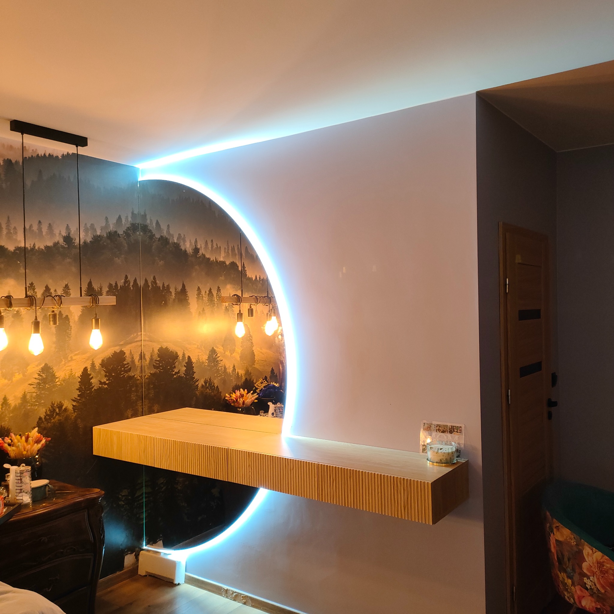 Nowoczesna aranżacja wnętrza z półką z ryflowanego drewna, podświetloną neonowym łukiem, na tle tapety z motywem leśnym i wiszącymi lampami żarówkowymi.