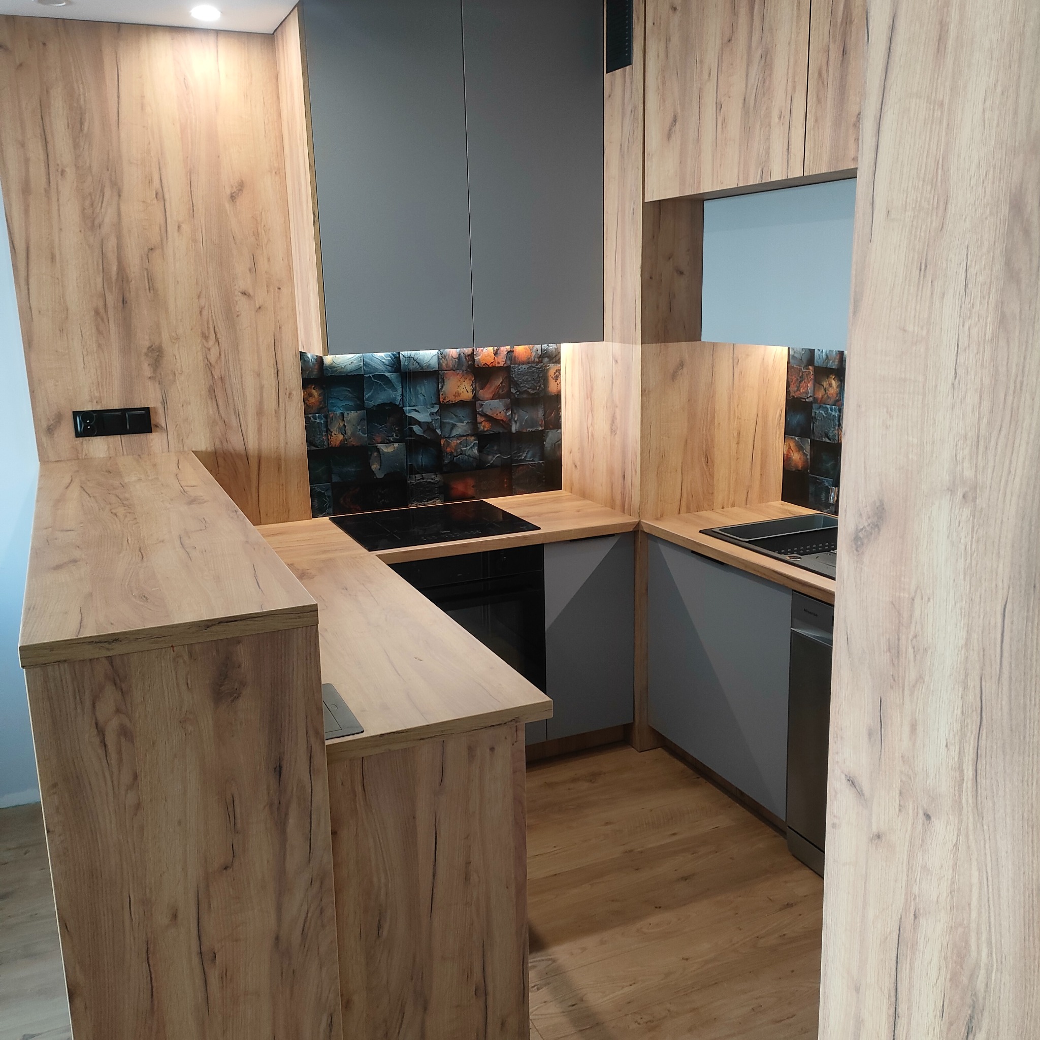 Nowoczesna kuchnia z drewnianymi frontami szafek i szarymi elementami, czarna płyta indukcyjna i piekarnik, oryginalna mozaika na ścianie nad blatem.