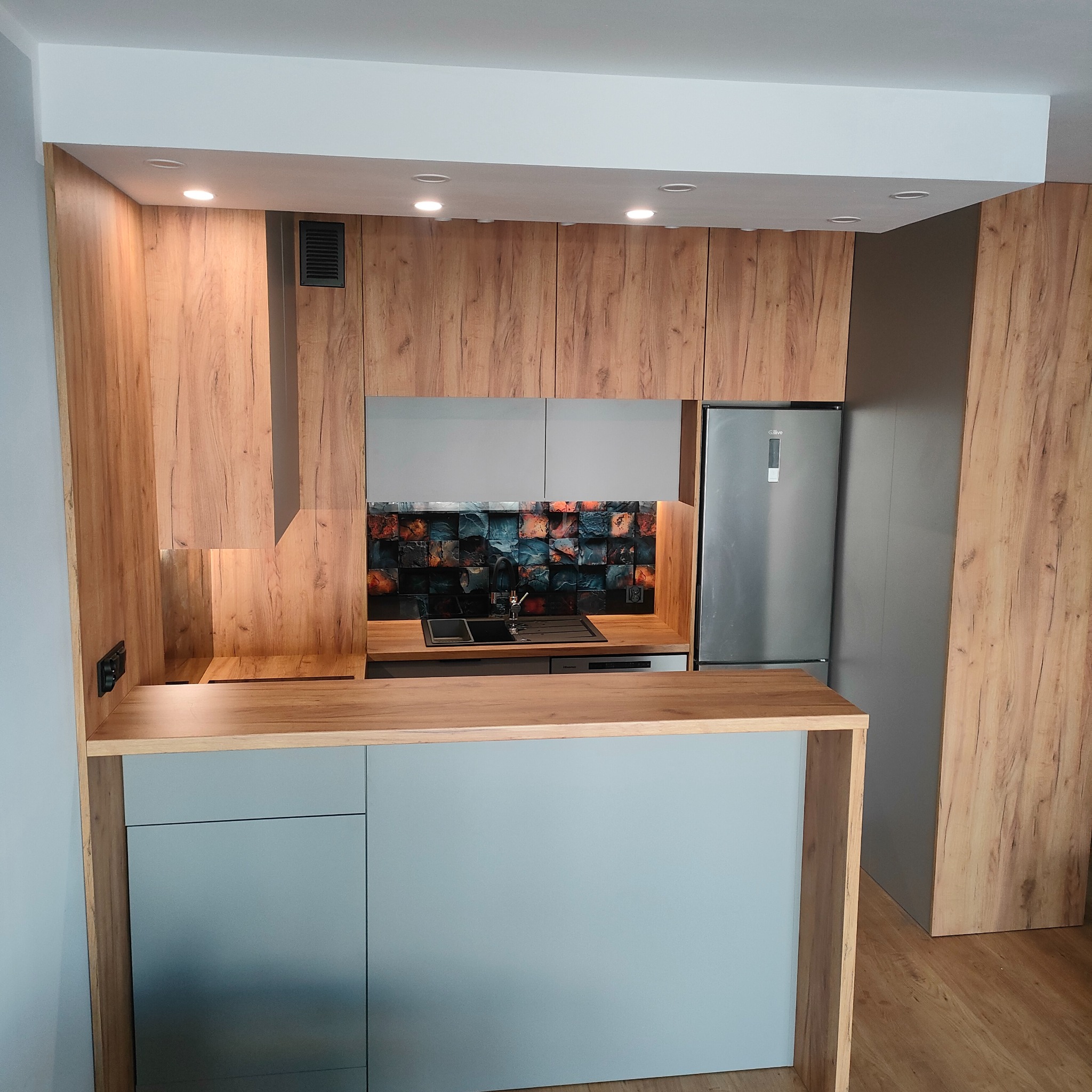 Nowoczesna zabudowa kuchenna z drewnianymi frontami, szarymi szafkami dolnymi i dekoracyjną mozaiką nad blatem roboczym, uzupełniona o srebrną lodówkę.