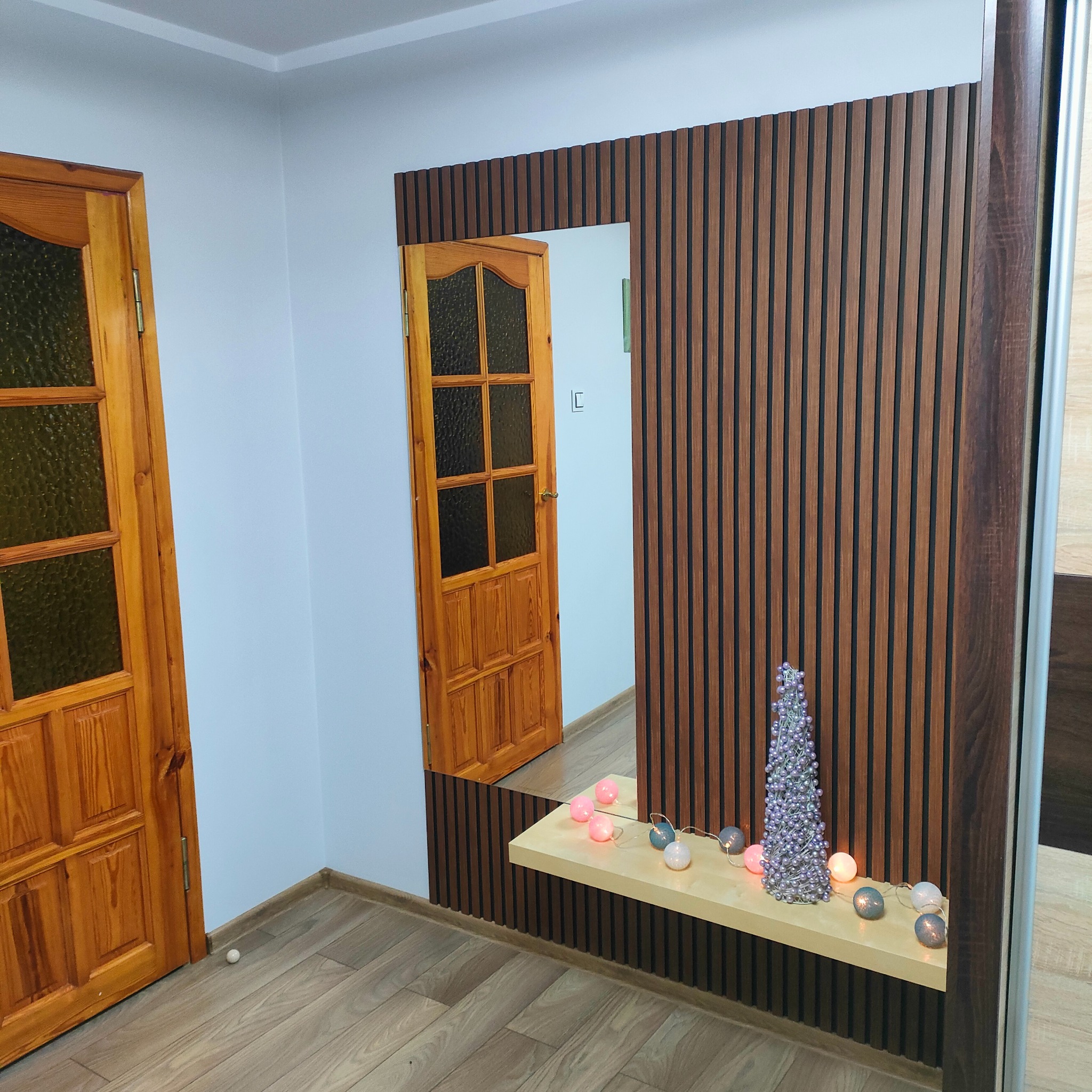 Nowoczesny przedpokój z drewnianą boazerią lamelową, lustrem wkomponowanym w boazerię, drewnianymi drzwiami z szybami i dekoracjami świątecznymi na półce
