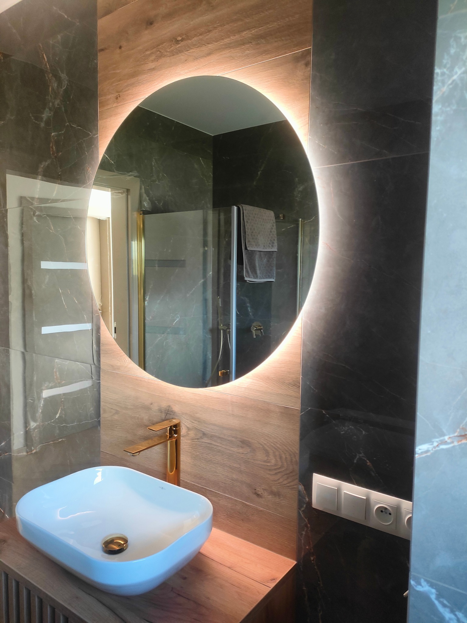 Nowoczesna łazienka z okrągłym lustrem podświetlanym LED, białą umywalką nablatową i złotą armaturą na tle płytek imitujących drewno i marmur.