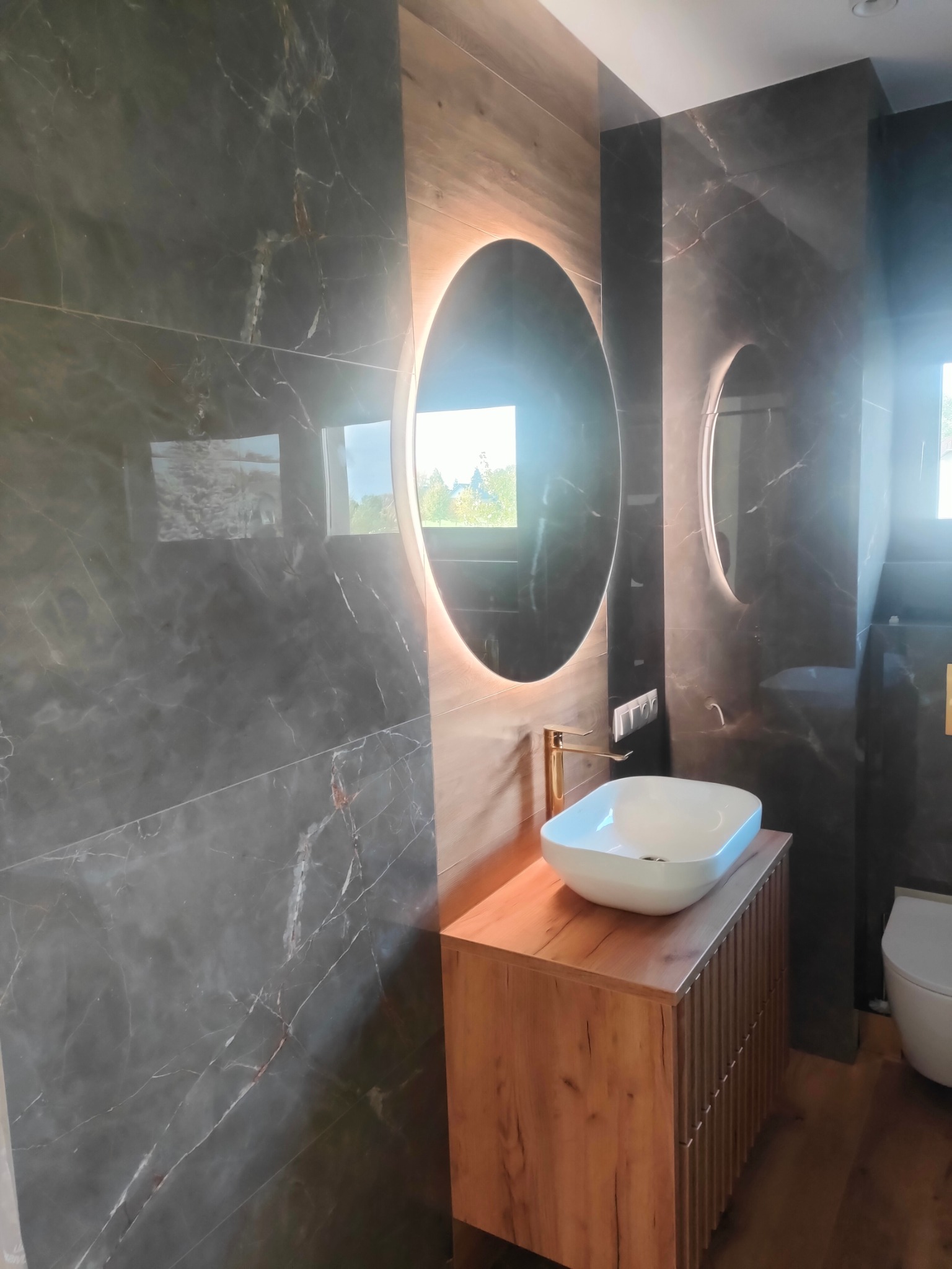 Nowoczesna łazienka z umywalką nablatową, okrągłym lustrem z podświetleniem LED i marmurowymi płytkami na ścianach.