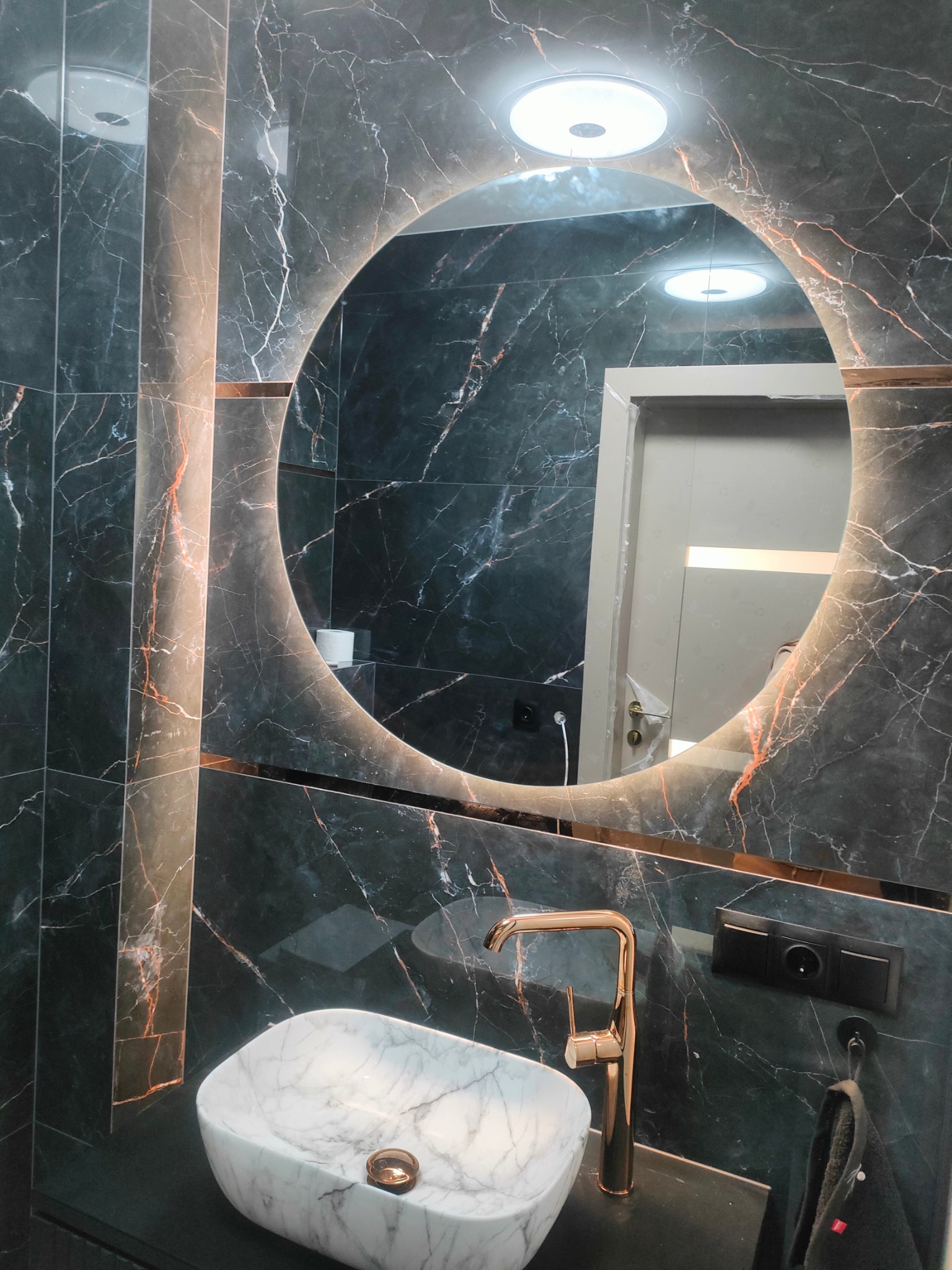 Nowoczesna łazienka z umywalką nablatową z białego marmuru, złotą baterią i okrągłym lustrem z podświetleniem LED na tle czarnych płytek marmurowych z brązowymi żyłkami.