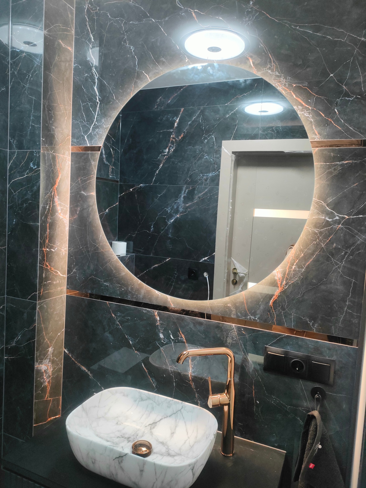 Nowoczesna łazienka z marmurowymi płytkami w odcieniach szarości i miedzianymi akcentami, okrągłe lustro z podświetleniem LED, umywalka nablatowa z marmuru Carrara i bateria w kolorze różowego złota.