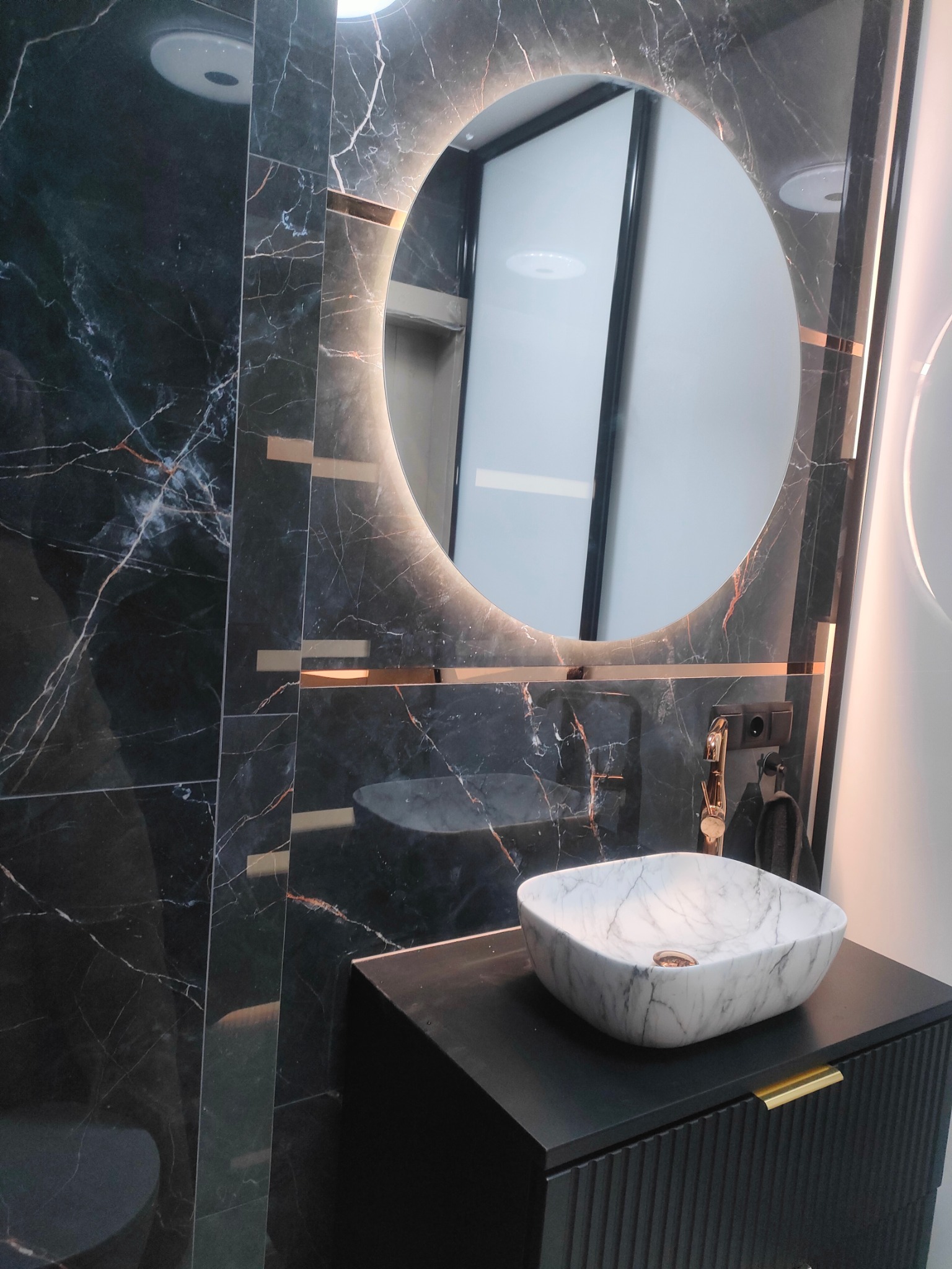 Nowoczesna łazienka z umywalką nablatową z marmuru, okrągłym lustrem z podświetleniem LED i czarnymi płytkami imitującymi marmur z złotymi akcentami.