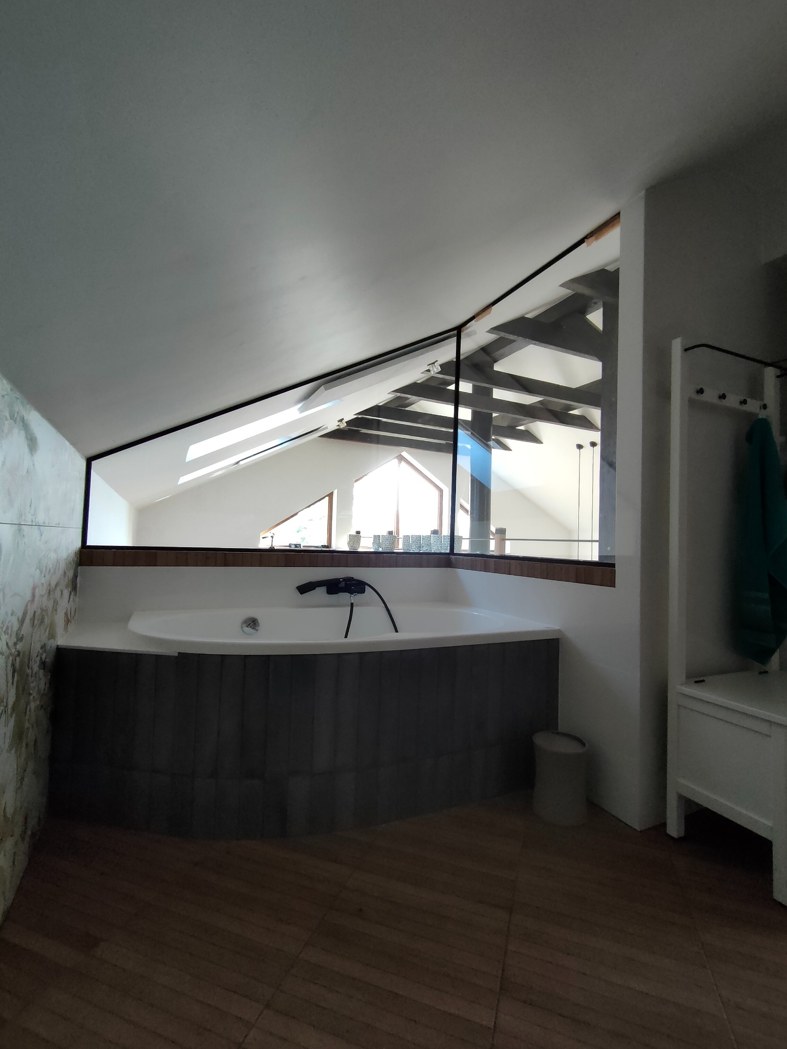 Nowoczesna łazienka na poddaszu z owalną wanną obudowaną szarą mozaiką, oddzielona od strychu szklaną ścianą z czarną ramą, widoczne drewniane elementy konstrukcyjne strychu i okno dachowe.