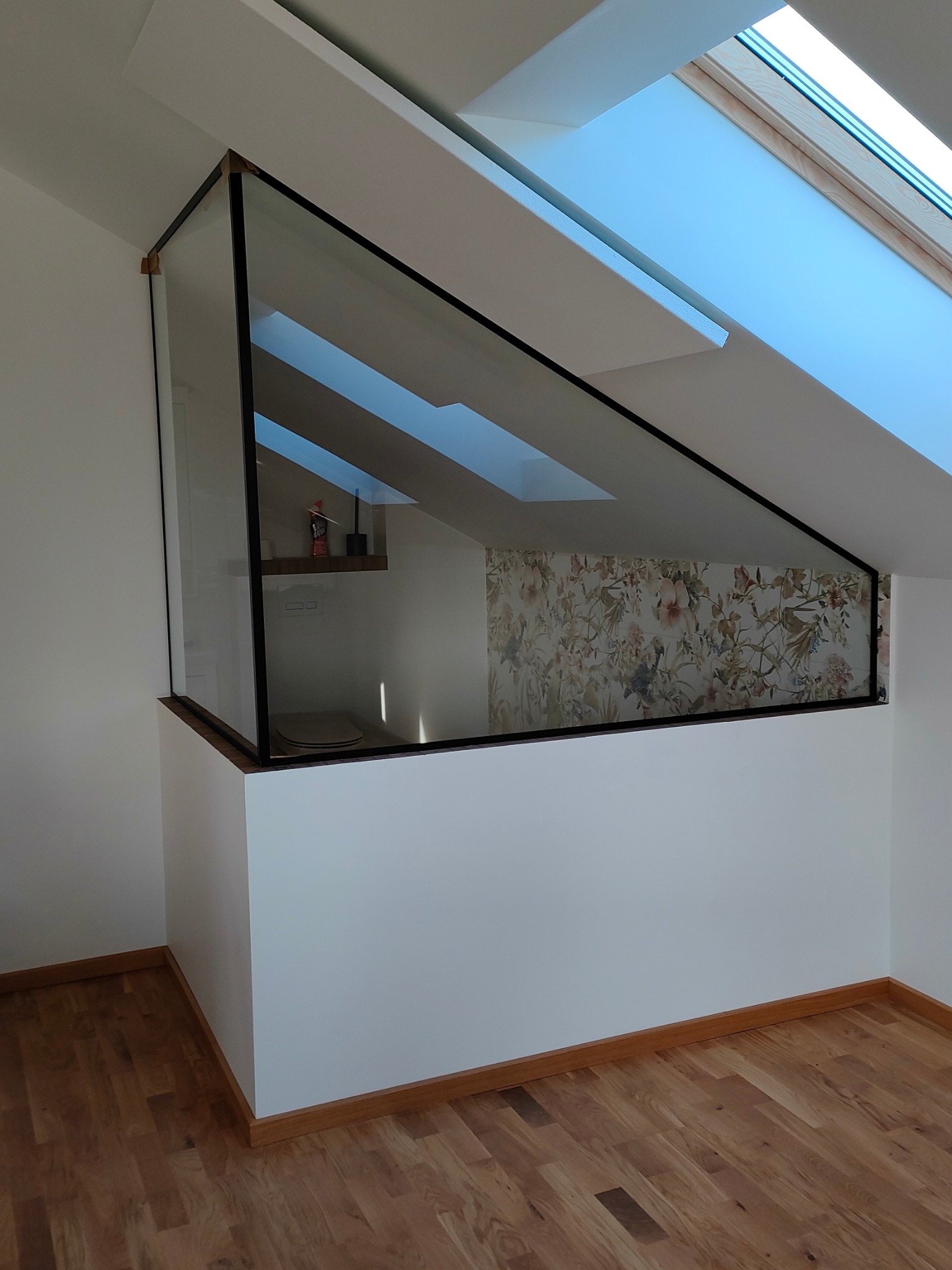 Szklana ścianka działowa z czarną ramą oddzielająca przestrzeń poddasza z oknem dachowym i tapetą w kwiaty, widok na drewnianą podłogę.