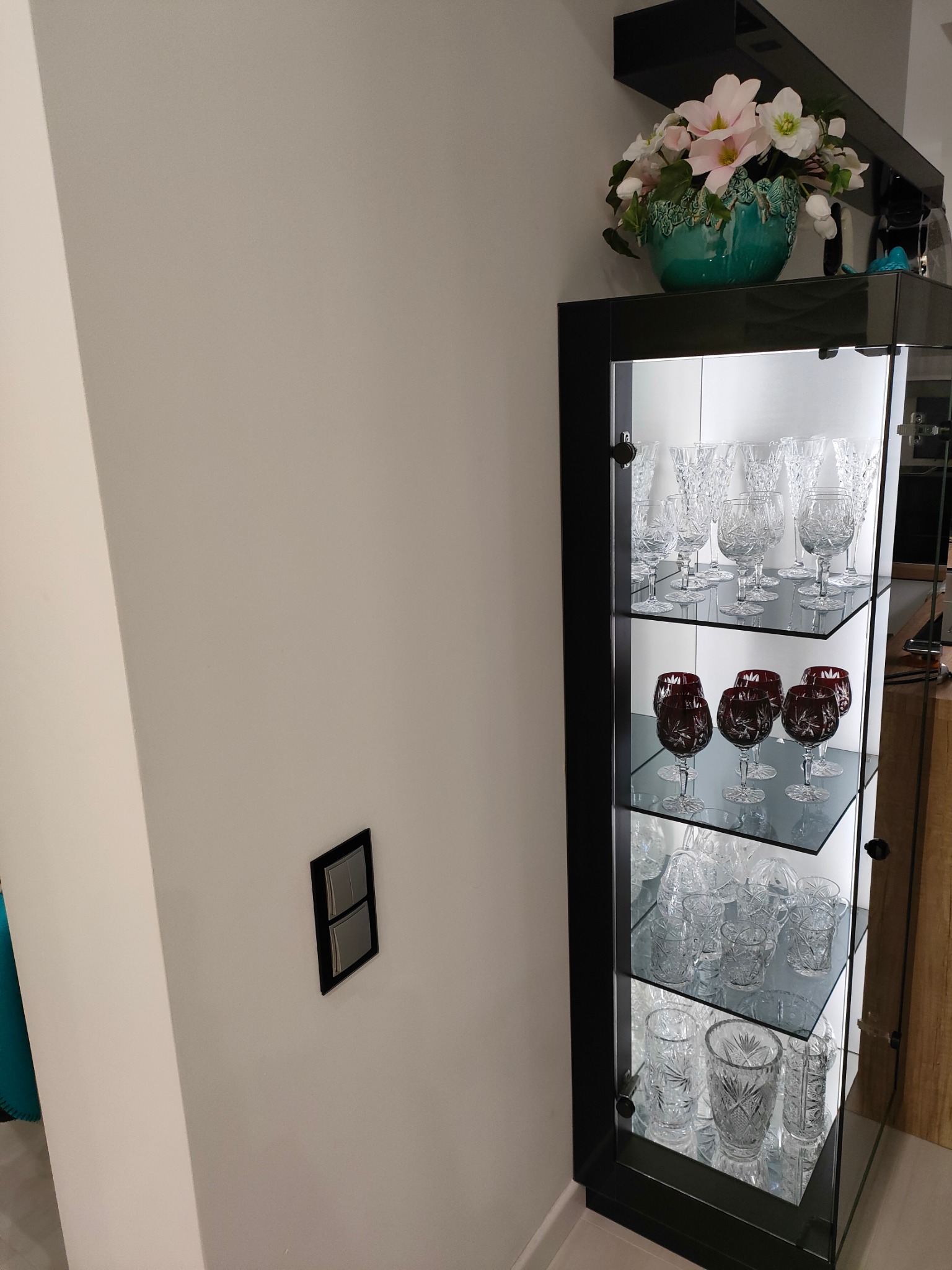 Szklana witryna w czarnej ramie, oświetlona od wewnątrz, prezentuje kolekcję kryształowych kieliszków i szklanek; na górze witryny dekoracyjny bukiet w turkusowym wazonie.