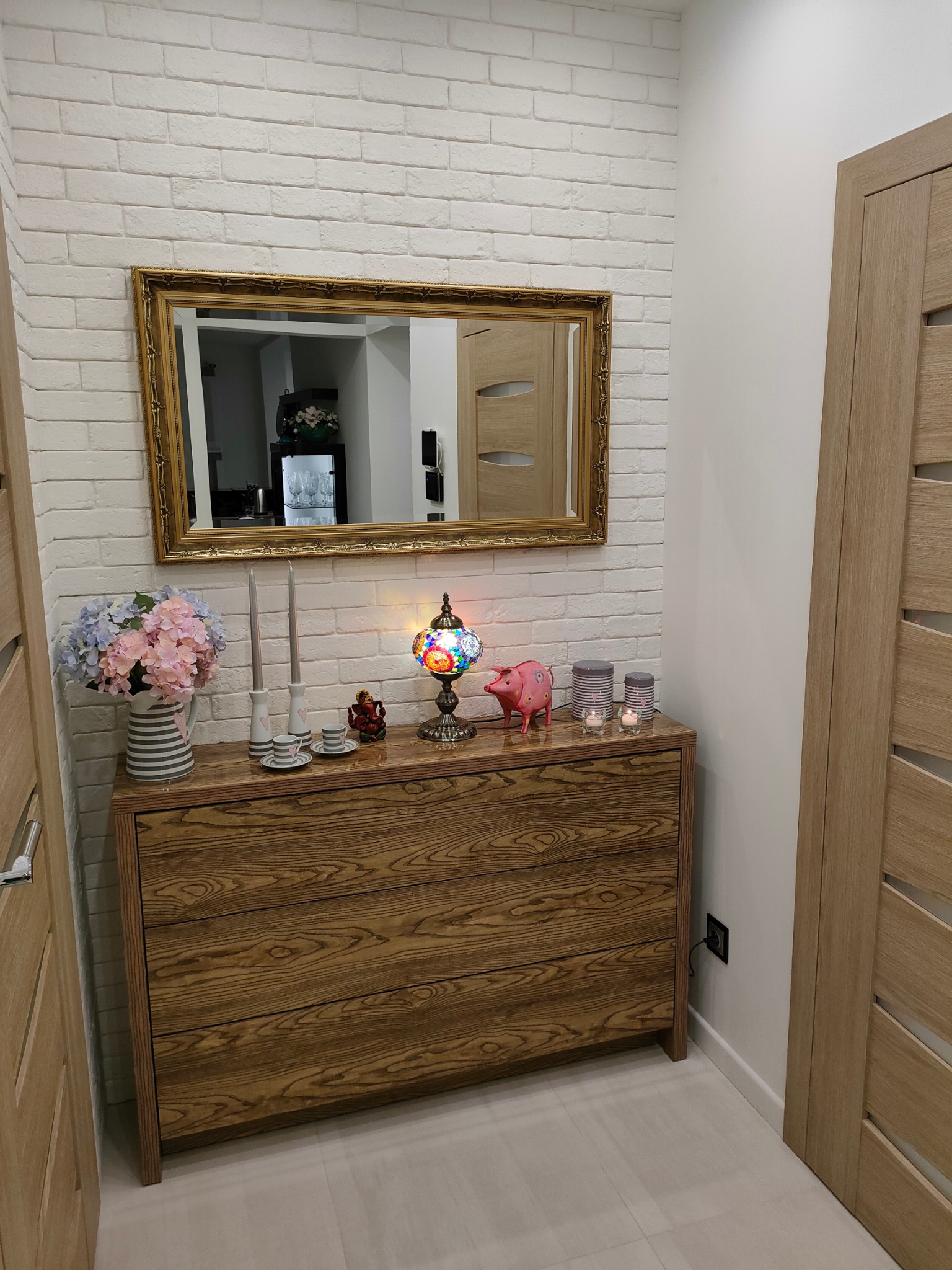 Dekoracyjny przedpokój z komodą z widocznym usłojeniem drewna, lustrem w złotej ramie na tle ściany z białej cegły, kwiatami w wazonie, ozdobną lampką i figurką świnki.
