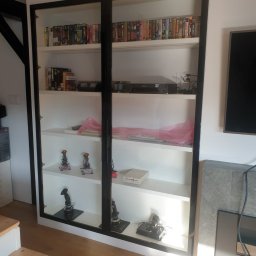 BEGI sp. z o.o. - Biała szafa z czarnymi ramami, wypełniona kasetami VHS i joystickami do gier, ustawiona w rogu pokoju obok schodów i kominka. Nietypowa ekspozycja kolekcji.