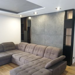 BEGI sp. z o.o. - Nowoczesny salon z szarą, modułową szafą wnękową z elementami drewna, oświetleniem punktowym i narożną sofą w odcieniach brązu na drewnianej podłodze.