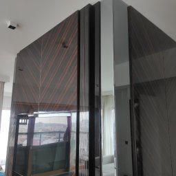 BEGI sp. z o.o. - Nowoczesna szafa wnękowa z forniru w geometryczny wzór, z lustrzanymi wstawkami, dzieląca przestrzeń pokoju z widokiem na miasto w tle.