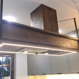 BEGI sp. z o.o. - Nowoczesny okap kuchenny z drewnianą obudową i oświetleniem LED, zawieszony nad szarymi szafkami kuchennymi. Widok z dołu, okno w tle.