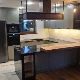 BEGI sp. z o.o. - Nowoczesna kuchnia z szarymi szafkami, marmurowym blatem i wyspą z czarnym blatem. Nad wyspą drewniana konstrukcja z oświetleniem. Widoczny sprzęt AGD.