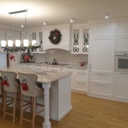 BEGI sp. z o.o. - Biała kuchnia na wymiar z wyspą i krzesłami barowymi, udekorowana świątecznym wieńcem i ozdobami. Klasyczne frezowane fronty i marmurowy blat.