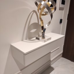 BEGI sp. z o.o. - Biała, wisząca szafka z szufladami, na niej dekoracyjna, metalowa rzeźba w odcieniach złota i srebra. Minimalistyczny design, marmurowa podłoga, nowoczesny interkom.