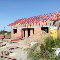 Wznoszona więźba dachowa z czerwonych belek na murowanym domu w stanie surowym otwartym, widoczna tablica informacyjna budowy i porozrzucane palety.