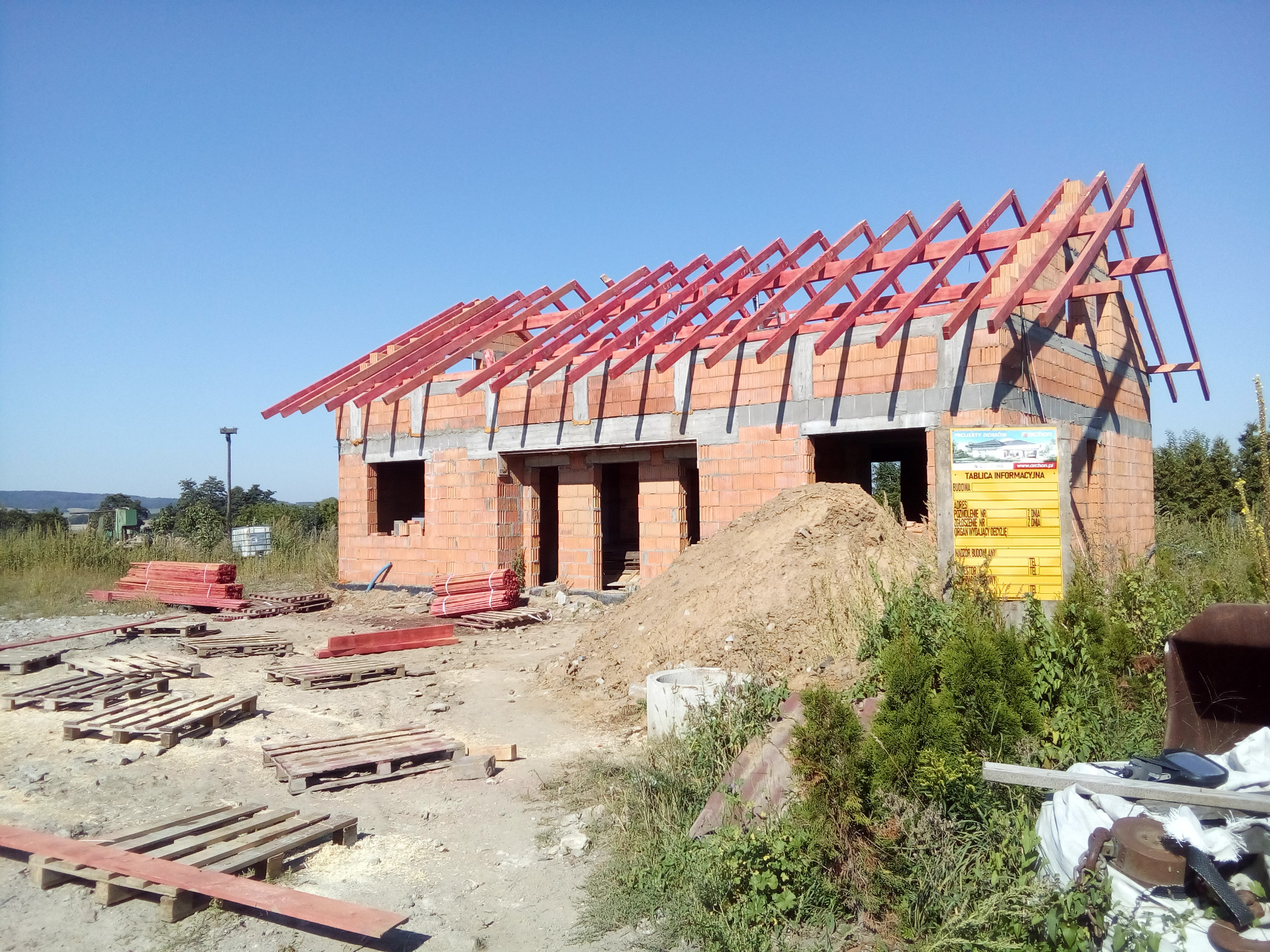 Wznoszona więźba dachowa z czerwonych belek na murowanym domu w stanie surowym otwartym, widoczna tablica informacyjna budowy i porozrzucane palety.