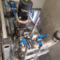 Welding technology Dmytro Kaniuka - Widok z dołu na przemysłową instalację pompową z silnikiem elektrycznym, rurociągami i zaworami z niebieskimi pokrętłami, umieszczoną na metalowej kratownicy, z fragmentem drabiny w kadrze.