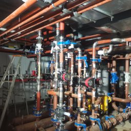 Welding technology Dmytro Kaniuka - Rozbudowana instalacja rur miedzianych z zaworami i pompami w kotłowni, widoczna drabina aluminiowa i szafy sterownicze na tle.