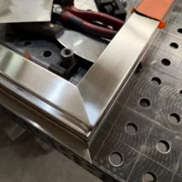 Nierdzewny narożnik ramy na stole spawalniczym z narzędziami. Precyzyjne połączenie elementów, widoczne detale szlifowania i wykończenia.