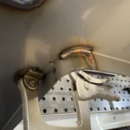 Metalowy element mocowany ściskiem spawalniczym, widoczny spaw z charakterystycznym zabarwieniem, tło stanowi stół spawalniczy z otworami.