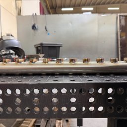 Welding technology Dmytro Kaniuka - Spawana rura ze stali nierdzewnej z precyzyjnymi spawami, leżąca na perforowanym stole spawalniczym w warsztacie. Widoczne ślady ciepła na spawach.