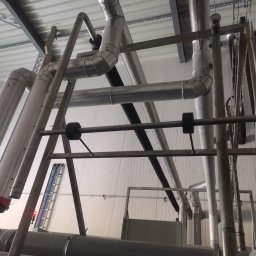 Welding technology Dmytro Kaniuka - Kompleksowa instalacja przemysłowa z rurami o różnych średnicach, mocowana do sufitu. Widoczne elementy mocujące i fragment drabiny w tle.