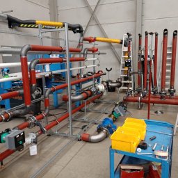 Welding technology Dmytro Kaniuka - Rozbudowana instalacja przemysłowa z rur w kolorach czerwonym, szarym i żółtym, zamontowana w hali. Widoczne liczne złącza, zawory i elementy mocujące. Na pierwszym planie wózek narzędziowy.