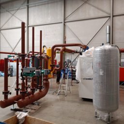 Welding technology Dmytro Kaniuka - Brązowe rury przemysłowe z zaworami i zielonymi regulatorami w hali produkcyjnej. W tle zbiorniki owinięte folią i drabina. Widoczny fragment pracownika.