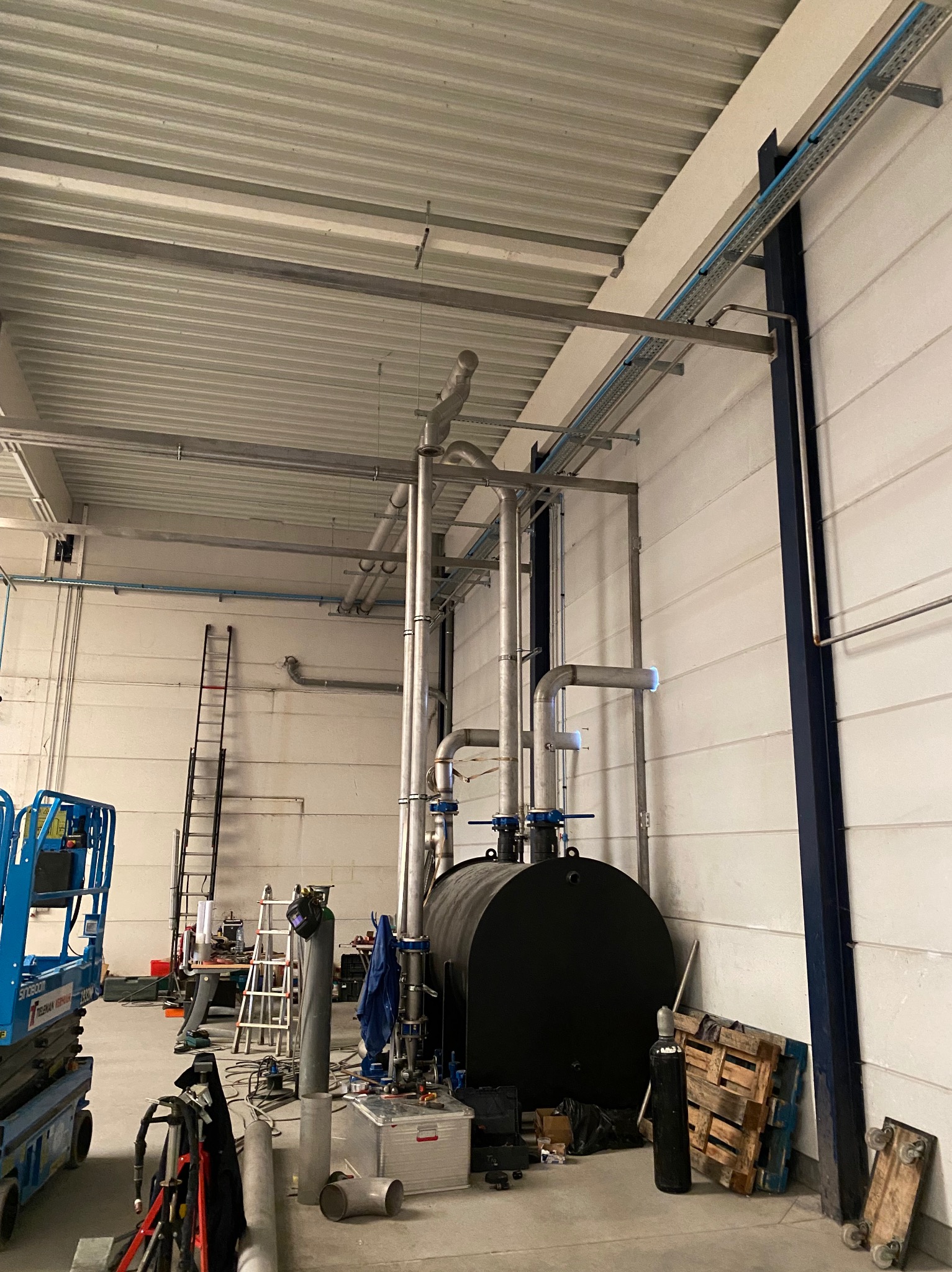 Montaż instalacji przemysłowej w hali: czarny zbiornik, rury stalowe i systemy mocowań pod sufitem, widoczny podnośnik koszowy i drabina.