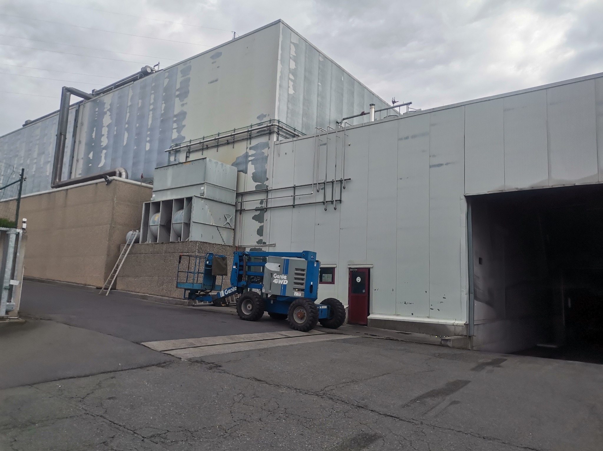 Niebieski podnośnik koszowy Genie 4WD zaparkowany przed budynkiem przemysłowym z panelami, obok drabina oparta o ścianę, widoczne instalacje wentylacyjne i rury na elewacji