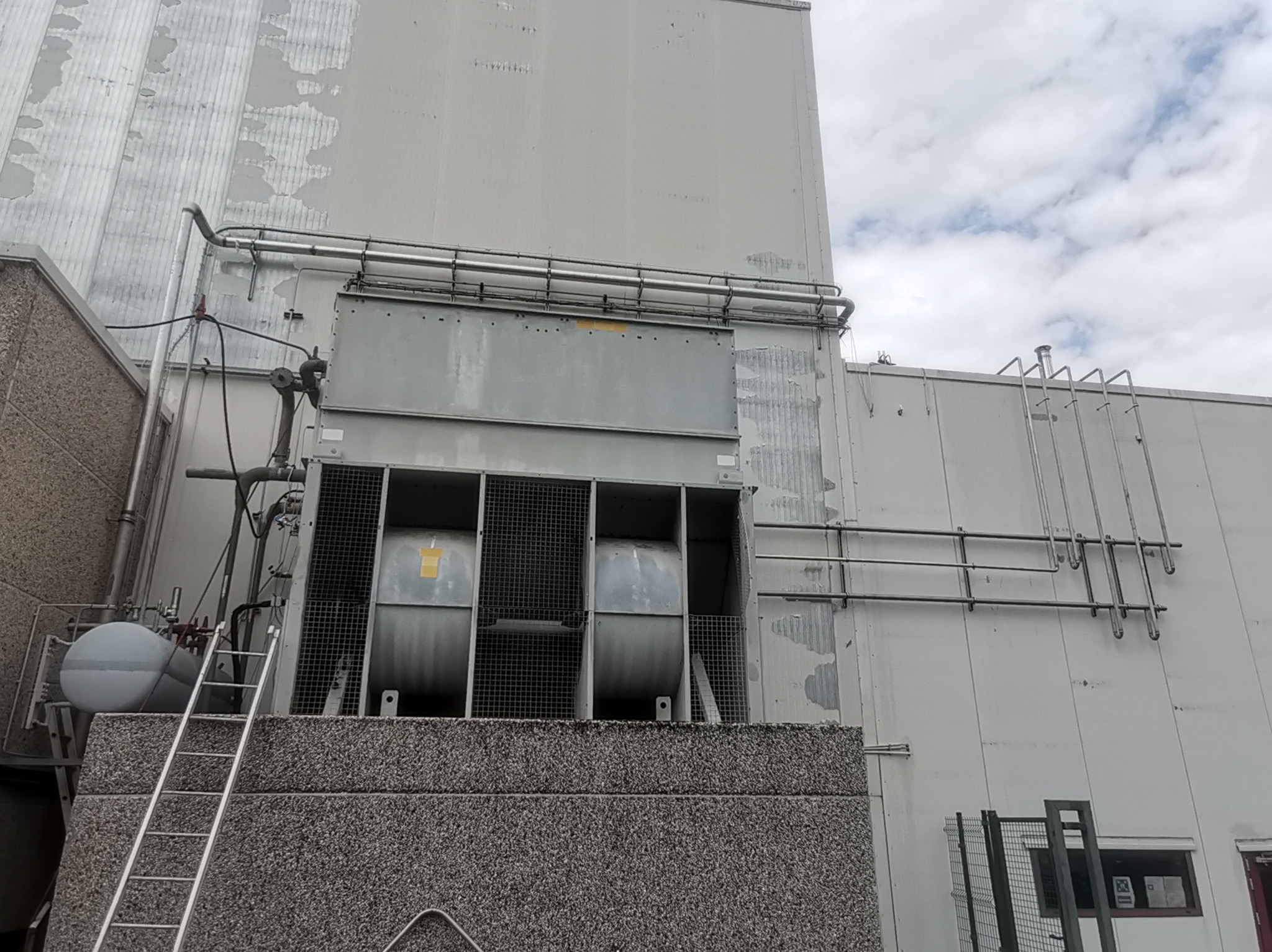 Zewnętrzna instalacja przemysłowa z wentylatorami, rurami i zbiornikiem, dostępna po metalowej drabinie, na tle szarej ściany budynku z widocznymi śladami użytkowania.