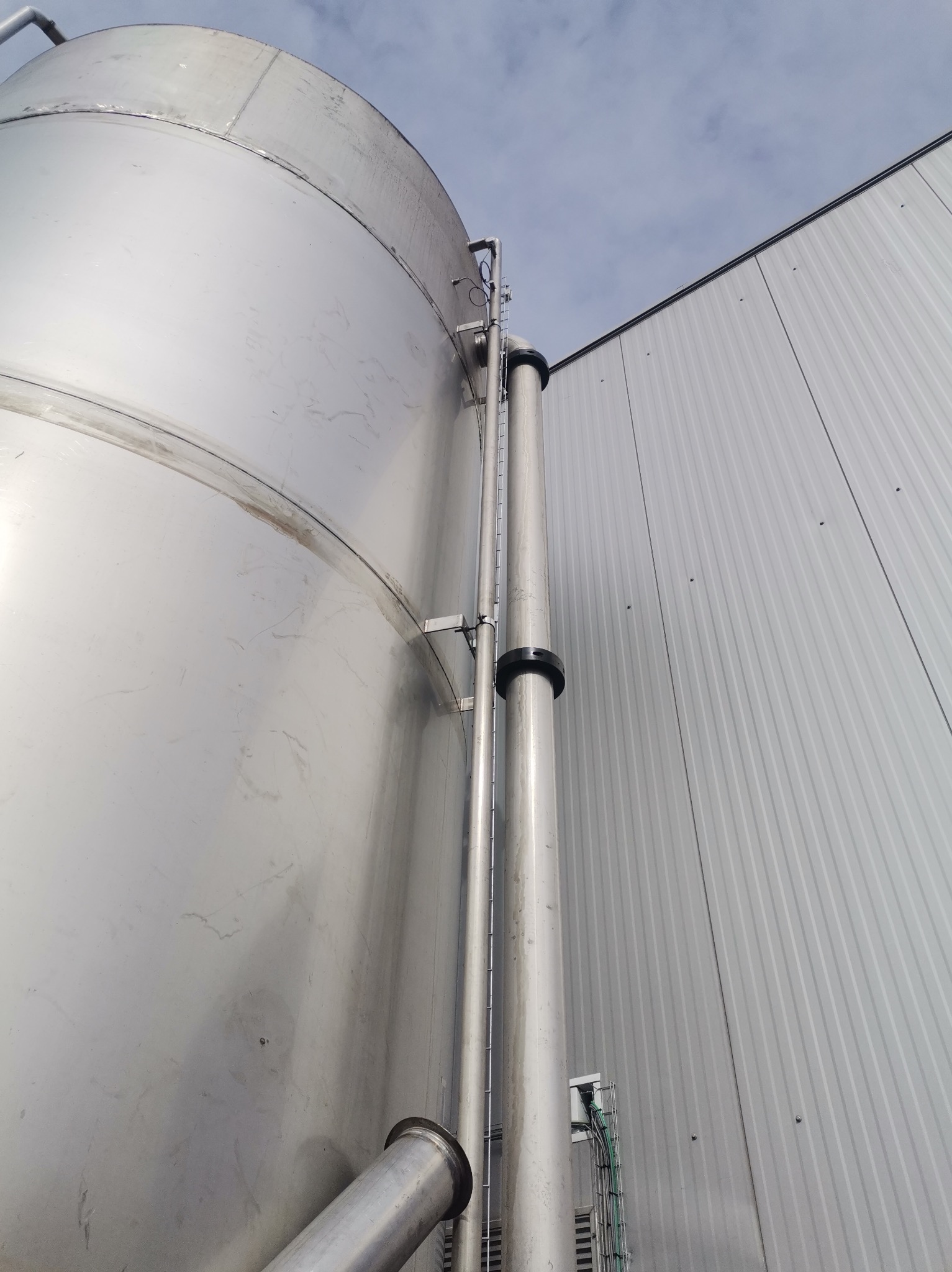 Zewnętrzny, stalowy zbiornik przemysłowy z rurami i drabinką, zamontowany przy hali o ścianach z blachy trapezowej, widziany z dołu pod kątem.