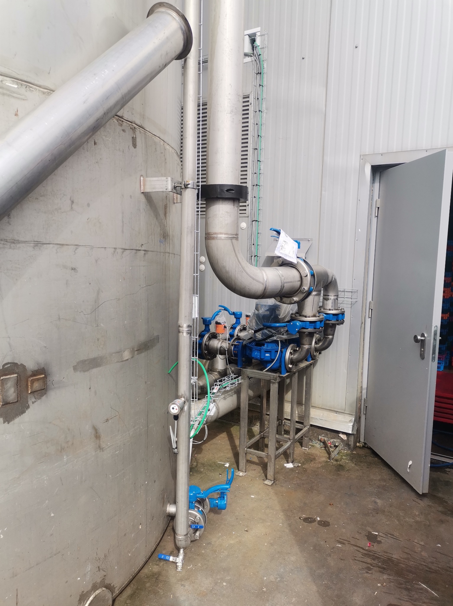 Srebrny zbiornik z systemem rur i niebieskimi pompami w hali przemysłowej, widoczna instalacja elektryczna i otwarte drzwi.