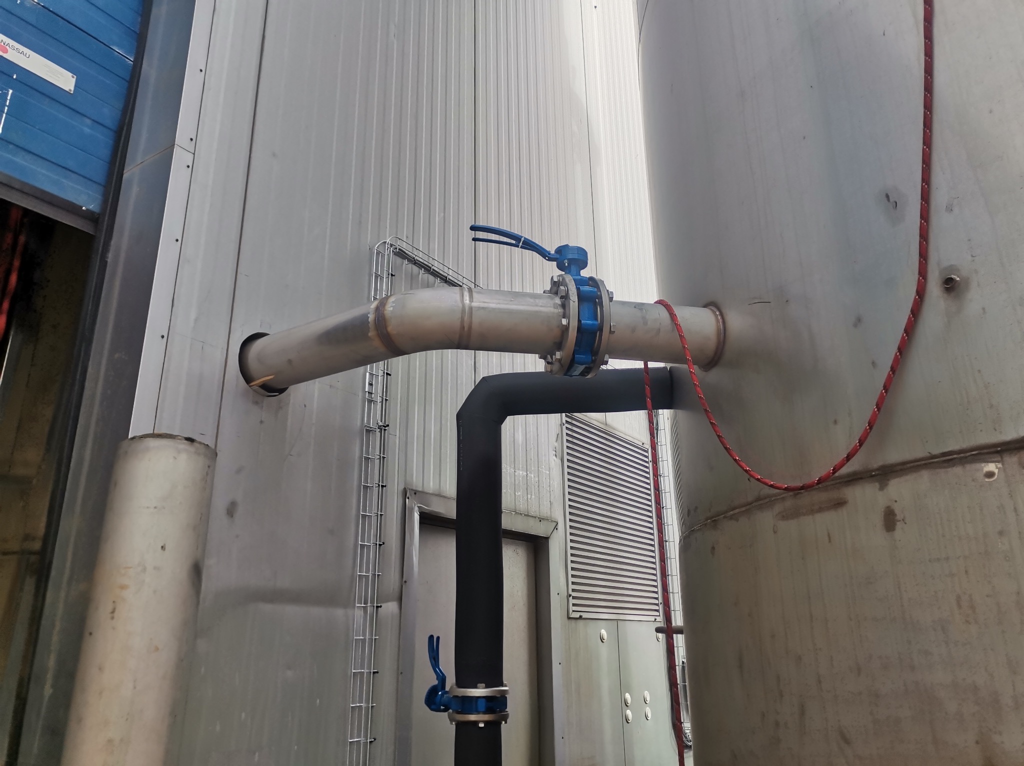 Instalacja przemysłowa z rurami i zaworami, widoczna część zbiornika, metalowa konstrukcja budynku w tle.