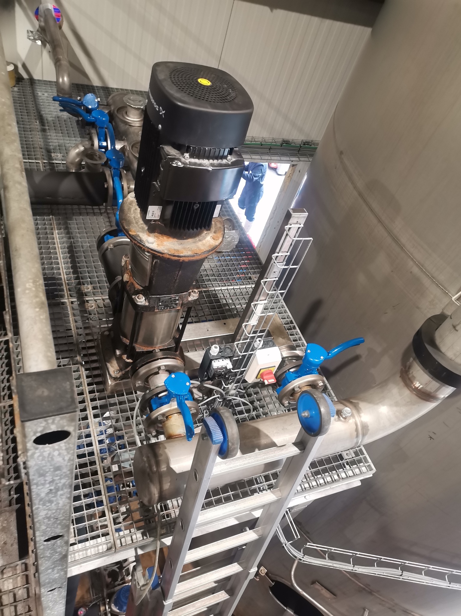 Widok z dołu na przemysłową instalację pompową z silnikiem elektrycznym, rurociągami i zaworami z niebieskimi pokrętłami, umieszczoną na metalowej kratownicy, z fragmentem drabiny w kadrze.