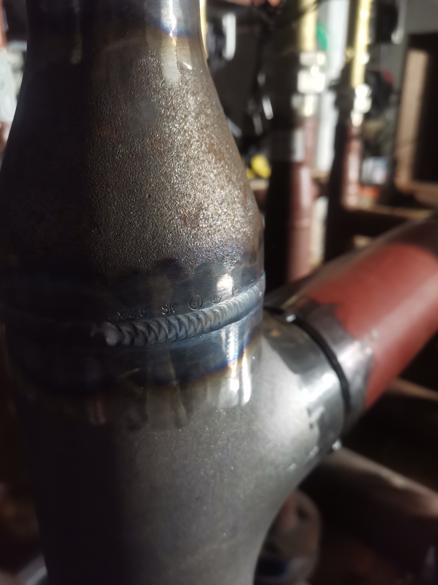 Połączenie spawane dwóch metalowych rur z widocznym szwem, z oznaczeniami 'X.2.6 SK' i symbolem graficznym, w tle zarys innych rur i elementów wyposażenia warsztatu.